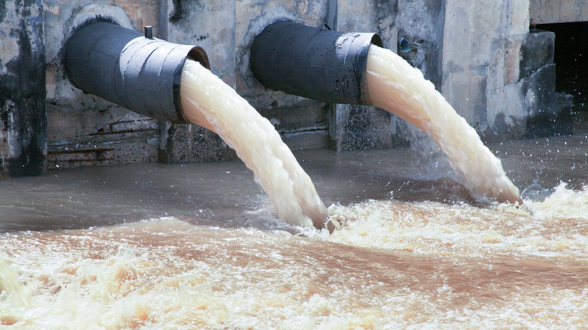 Zwei gro&szlig; dimensionierte Rohre leiten tr&uuml;bes Abwasser in ein offenes Gew&auml;sser; aufspritzende Str&ouml;mung und Sedimente sind deutlich erkennbar. Das Motiv unterstreicht die Bedeutung unabh&auml;ngiger Wasser- und Abwasseranalytik, Umweltmonitoring sowie normkonformer Pr&uuml;f- und Inspektionsleistungen f&uuml;r Betreiber in regulierten Branchen.