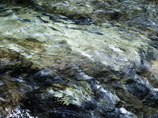 Nahaufnahme einer flach str&ouml;menden Wasseroberfl&auml;che mit sichtbaren Wirbeln, Lichtreflexen und dunklen, steinigen Bereichen darunter. Das Motiv steht f&uuml;r Umweltmonitoring und Wasseranalytik&mdash;ideal zur Illustration unabh&auml;ngiger Pr&uuml;f- und Laborleistungen f&uuml;r Trinkwasser-, Prozesswasser- und Gew&auml;sserqualit&auml;t in regulierten Anwendungen.